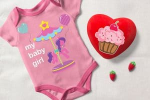 Top view of a pink baby bodysuit with balloons and pony rides illustration, a red heart shaped cushion with cupcakes on beige fabric.