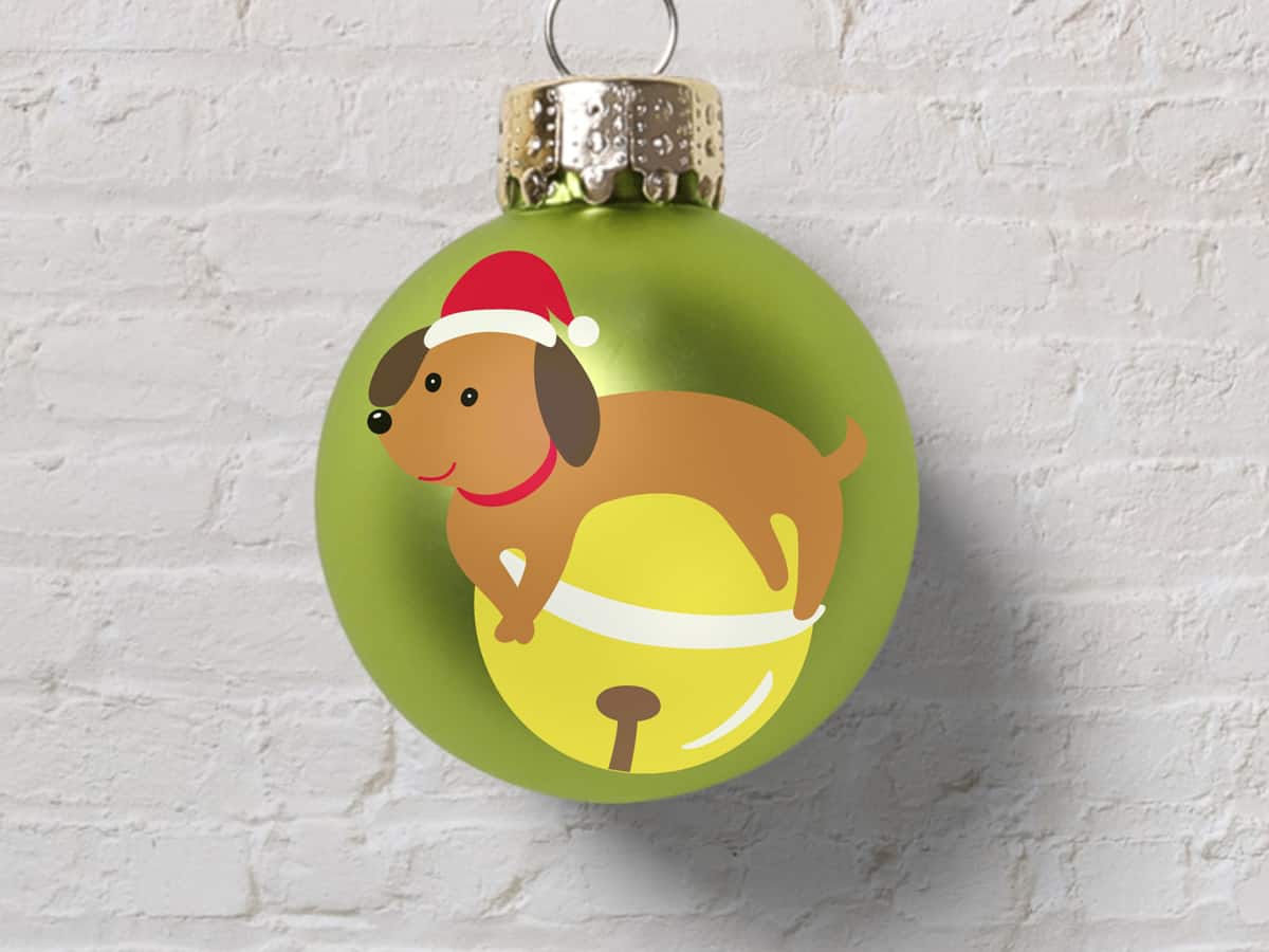 A green ornament with an illustration of a cute dog wearing a Santa hat sitting on a golden bell against a white wall
