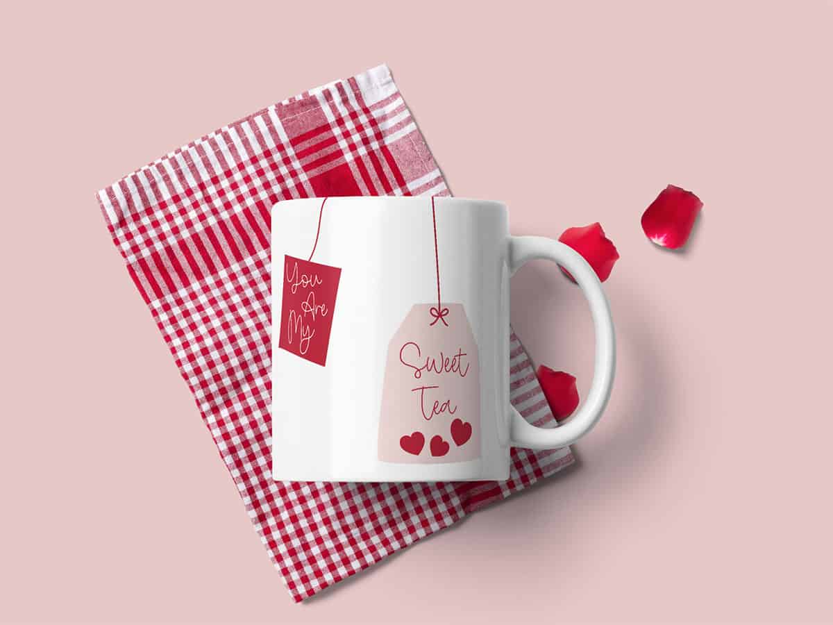 A white mug features an illustration of two tags with the text "You are my sweet tea" on a pink background and a red striped tea towel.