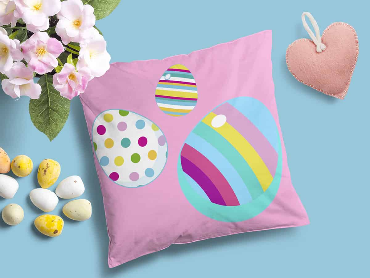 A top view of three Easter eggs on a pink pillow surrounded by flowers, small eggs and on a light blue background