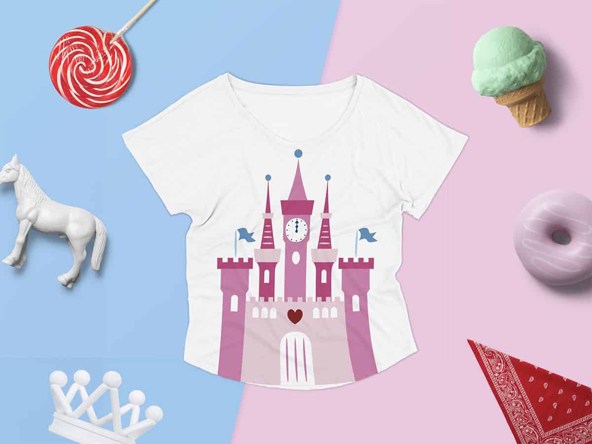 Kids' white T-shirt with a pink fairy-tale castle design, displayed with a lollipop, ice cream, and toys on a dual-tone blue and pink background.