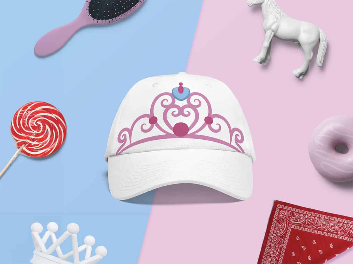 White baseball cap with a pink princess crown design, beside a red and white lollipop, pink hairbrush, white unicorn figurine, and a red bandana on a dual pink and blue background.
